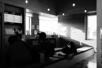 Several people are seated inside a waiting area with wooden walls and overhead lights. A digital board displaying information is fixed on the wall, and some decor and signage are visible. The area is illuminated by natural light entering through a window.