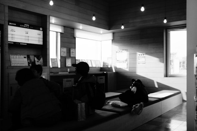 Several people are seated inside a waiting area with wooden walls and overhead lights. A digital board displaying information is fixed on the wall, and some decor and signage are visible. The area is illuminated by natural light entering through a window.