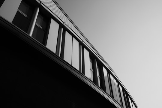 An architectural building facade with vertical pillars and multiple windows is captured from a low angle, emphasizing its geometric lines and modern design. The photo is presented in black and white, adding a dramatic and timeless effect.
