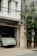 A pale green car is parked inside a garage under a residential building. Adjacent to the building is a telephone pole with numerous wires connected. There is also a shrub or small tree growing alongside the pole, partially covering it.