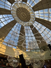 A glass-domed ceiling with geometric structures and a view of a modern building in the background. The dome is supported by metal frameworks, while spherical lighting fixtures and decorative elements hang from it.