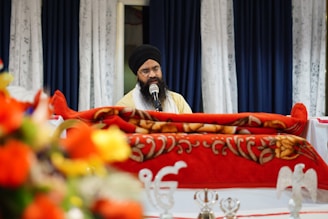 A man with a beard and turban is speaking into a microphone. He is seated behind a vibrant red, ornate cloth-covered structure. The background includes blue and white curtains, and a blurred foreground shows colorful floral arrangements.