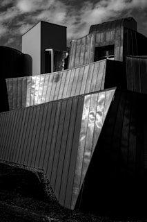 Architectural structure with modern and angular metal surfaces reflecting light, set against a cloudy sky. Offers a dramatic play of light and shadow across the geometric panels.