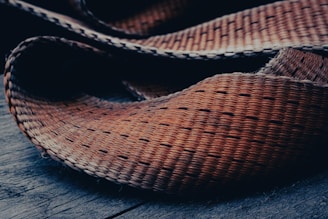 A close-up of a woven texture material, possibly a mat or basket, laid on a wooden surface. The fibers are tightly interlaced, showcasing a natural, hand-crafted feel.