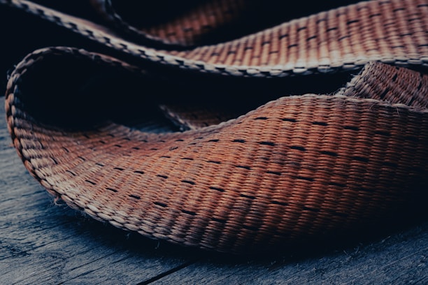 A close-up of a woven texture material, possibly a mat or basket, laid on a wooden surface. The fibers are tightly interlaced, showcasing a natural, hand-crafted feel.