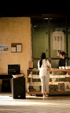 A person wearing a white top and jeans stands in front of a glass counter at a store. Behind the counter, another person appears to be assisting from the other side. The walls are aged and rustic, with one featuring a framed picture and another small notice. On the shelves under the counter, various items like books and records are displayed. A T-shirt hangs on the wall in a glass-enclosed space above the counter, and a large speaker is visible on the floor.