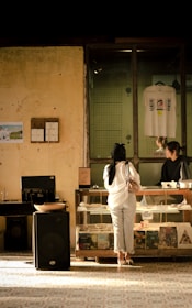 A person wearing a white top and jeans stands in front of a glass counter at a store. Behind the counter, another person appears to be assisting from the other side. The walls are aged and rustic, with one featuring a framed picture and another small notice. On the shelves under the counter, various items like books and records are displayed. A T-shirt hangs on the wall in a glass-enclosed space above the counter, and a large speaker is visible on the floor.