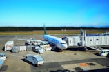 A commercial airplane is parked at an airport gate with multiple service vehicles surrounding it. There are cargo trucks, stair cars, and transportation carts handling luggage and other services. The aircraft appears to be preparing for departure or has just arrived. The sky is clear and the runway is visible in the background.