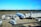 A commercial airplane is parked at an airport gate with multiple service vehicles surrounding it. There are cargo trucks, stair cars, and transportation carts handling luggage and other services. The aircraft appears to be preparing for departure or has just arrived. The sky is clear and the runway is visible in the background.