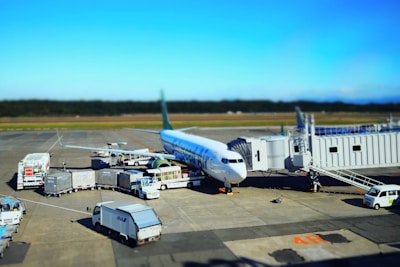 A commercial airplane is parked at an airport gate with multiple service vehicles surrounding it. There are cargo trucks, stair cars, and transportation carts handling luggage and other services. The aircraft appears to be preparing for departure or has just arrived. The sky is clear and the runway is visible in the background.