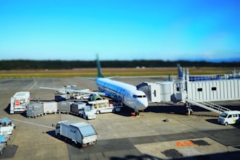 A commercial airplane is parked at an airport gate with multiple service vehicles surrounding it. There are cargo trucks, stair cars, and transportation carts handling luggage and other services. The aircraft appears to be preparing for departure or has just arrived. The sky is clear and the runway is visible in the background.