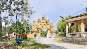 A gateway with ornate golden spires stands in the background, framed by tall trees. On the left side, there is a blue motorcycle parked on a grassy area. To the right, a traditional building with pink columns and a concrete balustrade is visible. The setting appears to be outdoors, with a clear sky enhancing the scene.