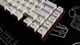 A close-up view of a mechanical keyboard featuring white keycaps and black legends on a dark surface. The keys display various navigational and functional labels like arrows, Home, Delete, Pg Up, and Pg Dn. The surrounding background includes faint outlines of retro gaming icons and technology symbols, contributing to a tech-themed aesthetic.