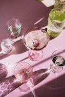 A collection of transparent glassware, including a variety of glasses and a bottle, is arranged on a pink surface. The light creates intricate patterns and reflections, with highlights and shadows accentuating the textures and contours of the glass.