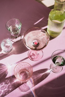 A collection of transparent glassware, including a variety of glasses and a bottle, is arranged on a pink surface. The light creates intricate patterns and reflections, with highlights and shadows accentuating the textures and contours of the glass.
