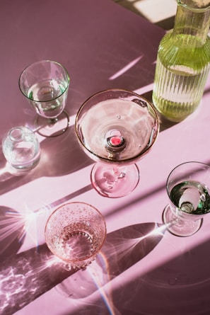 A collection of transparent glassware, including a variety of glasses and a bottle, is arranged on a pink surface. The light creates intricate patterns and reflections, with highlights and shadows accentuating the textures and contours of the glass.