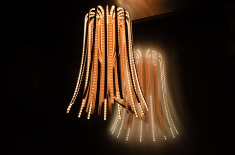 A unique and modern light fixture hangs from the ceiling, consisting of multiple long, flexible strips with embedded LED lights. The lights emit a warm glow against a dark background, creating an ambient atmosphere with the light reflecting on the wall.