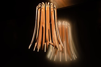 A unique and modern light fixture hangs from the ceiling, consisting of multiple long, flexible strips with embedded LED lights. The lights emit a warm glow against a dark background, creating an ambient atmosphere with the light reflecting on the wall.