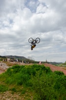 A cyclist performs an impressive mid-air flip on a dirt track surrounded by green vegetation, with a cloudy sky and distant hills forming the backdrop. A group of spectators observes the stunt from below.