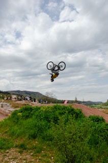 A cyclist performs an impressive mid-air flip on a dirt track surrounded by green vegetation, with a cloudy sky and distant hills forming the backdrop. A group of spectators observes the stunt from below.