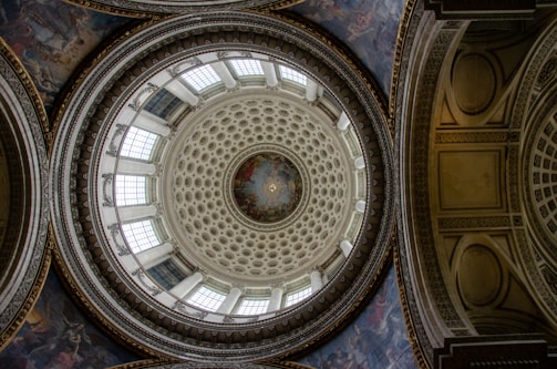 An intricately designed dome with ornate patterns and detailed artwork. The circular pattern features layers of designs with a central circular mural surrounded by geometric shapes. Large windows encircle the dome, allowing natural light to illuminate the space. The architectural details include ornate moldings and decorative plasterwork.