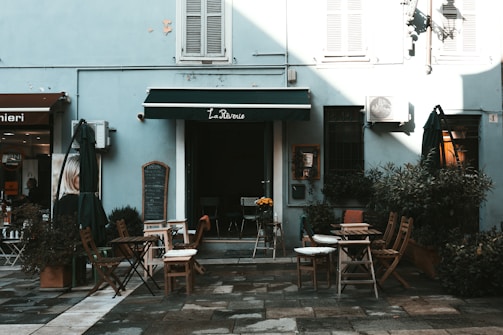 A small outdoor café with several wooden tables and chairs arranged on a paved area. The café has a blue facade with a dark green awning labeled 'La Reverie'. Various plants in pots are placed around the seating area, and outdoor umbrellas are folded next to some tables. The café is situated next to another store with a sign in Italian.