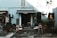 A small outdoor café with several wooden tables and chairs arranged on a paved area. The café has a blue facade with a dark green awning labeled 'La Reverie'. Various plants in pots are placed around the seating area, and outdoor umbrellas are folded next to some tables. The café is situated next to another store with a sign in Italian.