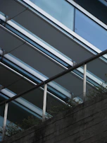 A modern architectural structure is captured, featuring parallel metal beams and glass panels creating a sleek, angular design. The lower part of the image shows a textured concrete surface with a metal railing, where greenery peeks through the gaps, adding a natural element to the urban setting.