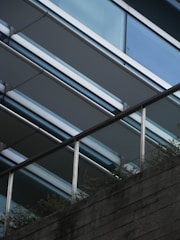 A modern architectural structure is captured, featuring parallel metal beams and glass panels creating a sleek, angular design. The lower part of the image shows a textured concrete surface with a metal railing, where greenery peeks through the gaps, adding a natural element to the urban setting.
