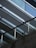 A modern architectural structure is captured, featuring parallel metal beams and glass panels creating a sleek, angular design. The lower part of the image shows a textured concrete surface with a metal railing, where greenery peeks through the gaps, adding a natural element to the urban setting.