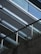 A modern architectural structure is captured, featuring parallel metal beams and glass panels creating a sleek, angular design. The lower part of the image shows a textured concrete surface with a metal railing, where greenery peeks through the gaps, adding a natural element to the urban setting.