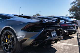 A lineup of exotic sports cars featuring sleek designs and shiny surfaces, parked in an outdoor lot under a clear blue sky. The focus is on the rear of the vehicles, showcasing their aerodynamic curves and detailing.