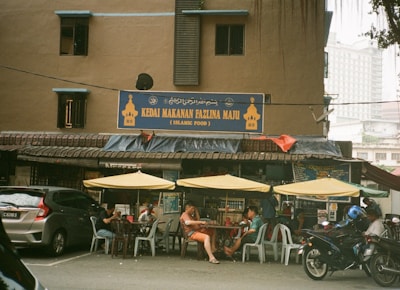 A street-side Islamic food restaurant with a sign displaying 'Kedai Makanan Fazlina Maju' above the entrance. The exterior features a brown building with small windows and a slanted roof. Several yellow umbrellas provide shade for customers seated at plastic tables and chairs outside. Various vehicles, including a car and a motorcycle, are parked in front of the restaurant.