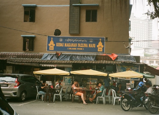 A street-side Islamic food restaurant with a sign displaying 'Kedai Makanan Fazlina Maju' above the entrance. The exterior features a brown building with small windows and a slanted roof. Several yellow umbrellas provide shade for customers seated at plastic tables and chairs outside. Various vehicles, including a car and a motorcycle, are parked in front of the restaurant.
