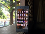 A smartphone displaying its home screen with various app icons is placed on a wooden surface. The background shows a dimly lit interior with a window or lattice through which sunlight filters, revealing greenery outside.