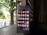 A smartphone displaying its home screen with various app icons is placed on a wooden surface. The background shows a dimly lit interior with a window or lattice through which sunlight filters, revealing greenery outside.