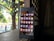 A smartphone displaying its home screen with various app icons is placed on a wooden surface. The background shows a dimly lit interior with a window or lattice through which sunlight filters, revealing greenery outside.