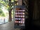 A smartphone displaying its home screen with various app icons is placed on a wooden surface. The background shows a dimly lit interior with a window or lattice through which sunlight filters, revealing greenery outside.