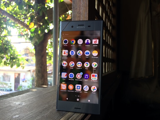 A smartphone displaying its home screen with various app icons is placed on a wooden surface. The background shows a dimly lit interior with a window or lattice through which sunlight filters, revealing greenery outside.