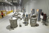 A large, industrial facility with multiple cylindrical metal tanks equipped with various pipes and valves. Control panels with lit indicators are mounted on the walls. A person in a reflective vest walks alongside a row of yellow safety railings.
