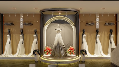 An elegant store display features multiple mannequins dressed in beautiful white gowns, with a central spotlight on a mannequin wearing a sparkling gown. The background includes racks with hangers, and there are flower arrangements at the base of the display. The ambiance is sophisticated with warm lighting.