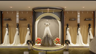 An elegant store display features multiple mannequins dressed in beautiful white gowns, with a central spotlight on a mannequin wearing a sparkling gown. The background includes racks with hangers, and there are flower arrangements at the base of the display. The ambiance is sophisticated with warm lighting.