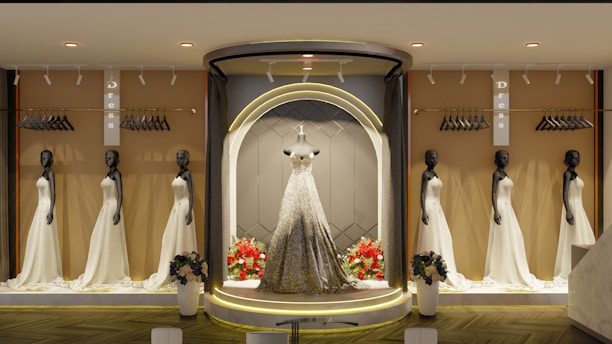 An elegant store display features multiple mannequins dressed in beautiful white gowns, with a central spotlight on a mannequin wearing a sparkling gown. The background includes racks with hangers, and there are flower arrangements at the base of the display. The ambiance is sophisticated with warm lighting.