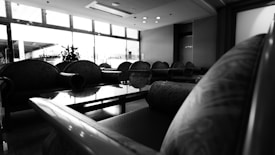 A black and white image of an interior space featuring multiple cushioned seats arranged in a lounge area with a glass coffee table. Large windows allow natural light to fill the space, creating a contrast of shadows and highlights.