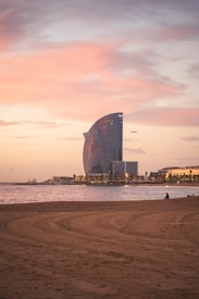 A beachfront view featuring a large, sail-shaped modern building against a soft pink and orange sunset sky. The scene includes a sandy beach with visible tire marks and a calm sea reflecting the sky's colors. Several birds and an airplane are flying in the background, and a few people can be seen sitting on the beach.