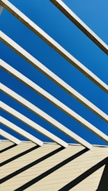 Wooden beams are arranged in a slanted pattern, casting shadows on a light-colored surface beneath them. The beams contrast sharply with the clear blue sky in the background.