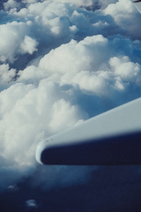 An airplane wing is visible against a backdrop of fluffy white clouds, with the sky providing a soft blue hue. The wing is slightly blurred, emphasizing the height and vastness of the scene.