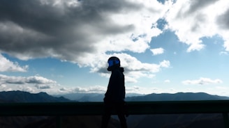 A person wearing a helmet with a reflective visor stands silhouetted against a backdrop of vast mountains and a dramatic sky filled with clouds. The contrasting light creates a striking balance between the dark silhouette and the bright sky.