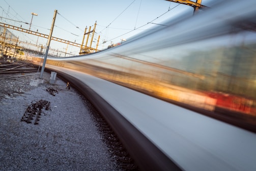 A high-speed train blurs past on a railway track, surrounded by a network of electric cables and infrastructural elements. The motion of the train creates a dynamic visual effect, contrasting with the static elements of the railway environment, such as tracks, overhead wires, and support poles.