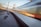 A high-speed train blurs past on a railway track, surrounded by a network of electric cables and infrastructural elements. The motion of the train creates a dynamic visual effect, contrasting with the static elements of the railway environment, such as tracks, overhead wires, and support poles.